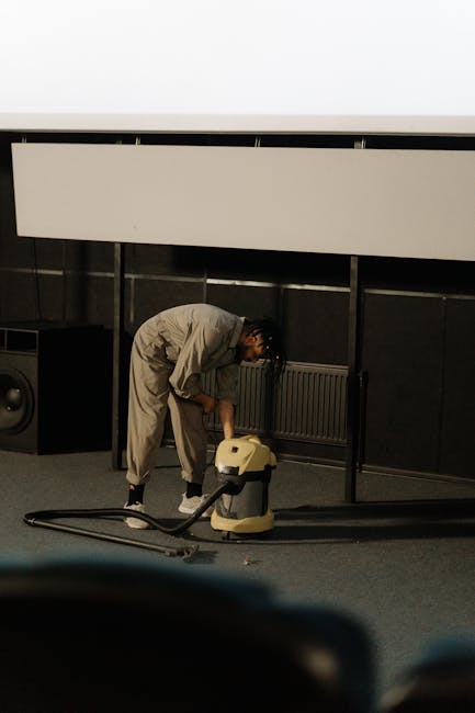 A woman using a vacuum cleaner to deep clean and remove dust from an oriental-style area rug in a living room. The room features wooden flooring and a brown leather sofa, with a wooden console table a