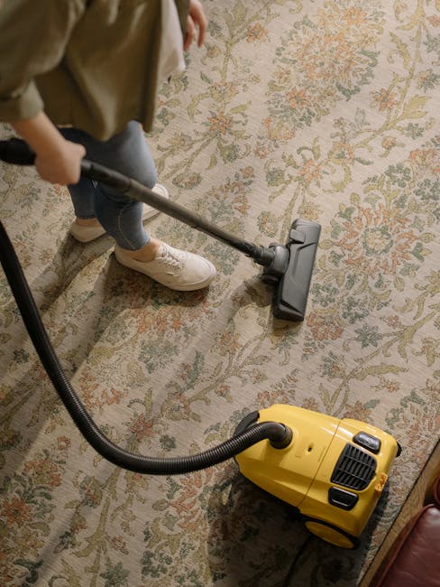 A person vacuuming a large, ornate area rug with a yellow canister vacuum cleaner in a well-lit room. The individual is wearing light-colored sneakers and casual clothing, holding the vacuum's wand at